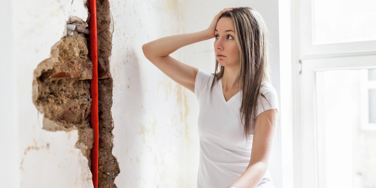 Frau steht besorgt vor einer geöffneten Innenwand mit freigelegtem Schaden und sichtbarer Leitung.